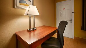A hotel room with a wooden desk, a black mesh chair, a white door, and a framed picture on the wall.