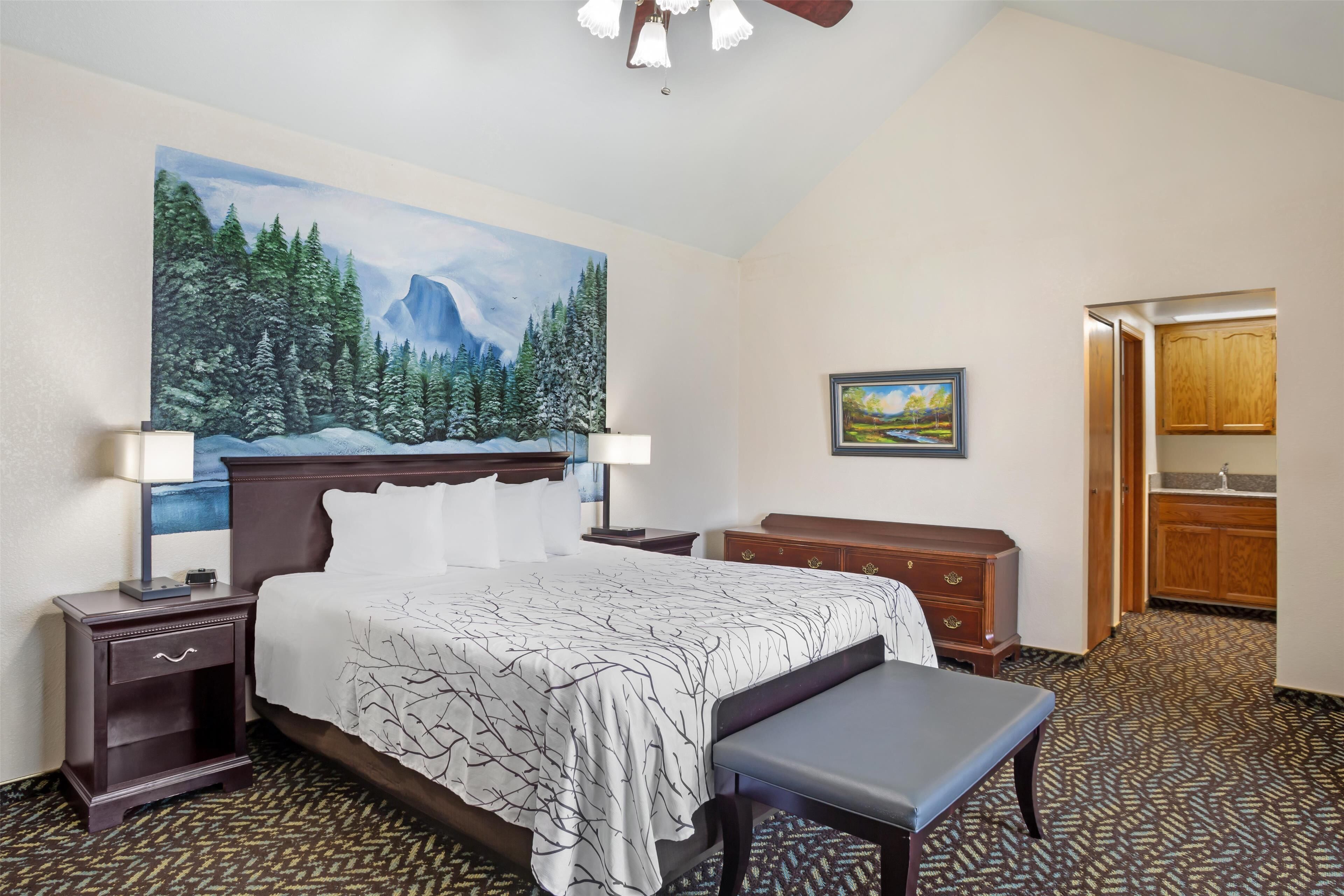 A hotel room with a large bed, a bench, a nightstand, a painting, and a view of the kitchen.