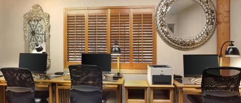 A hotel room with a wooden desk, two office chairs, two monitors, a printer, a lamp, a decorative mirror, and a window with blinds.