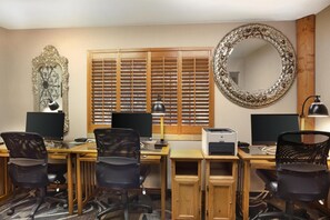 A hotel room with a wooden desk, two office chairs, two monitors, a printer, a lamp, a decorative mirror, and a window with blinds.