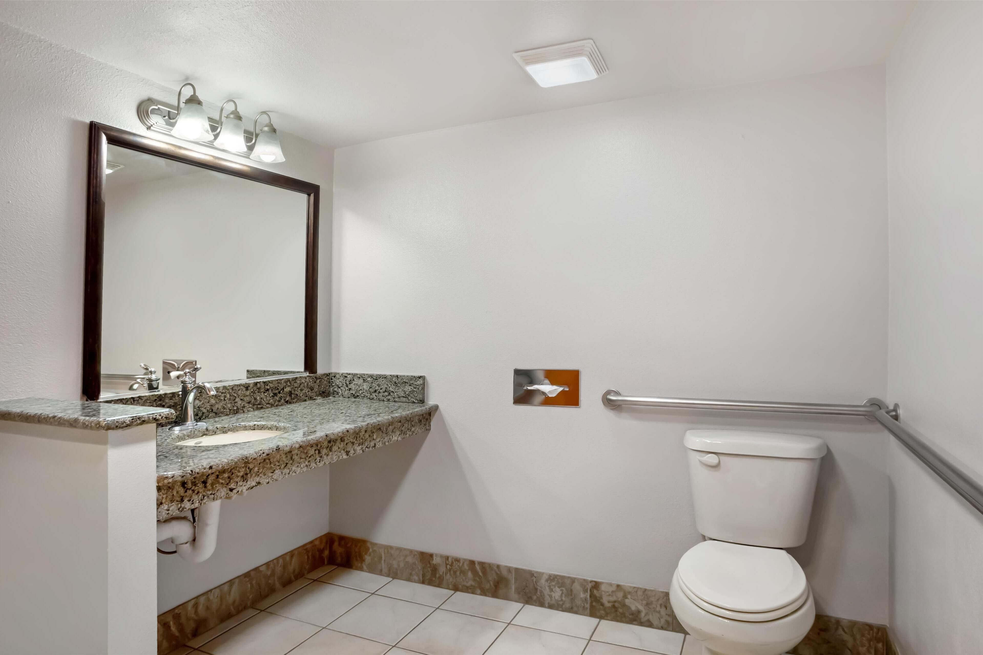Un baño con lavamanos de encimera de granito, un espejo grande, un inodoro blanco y un pasamanos.