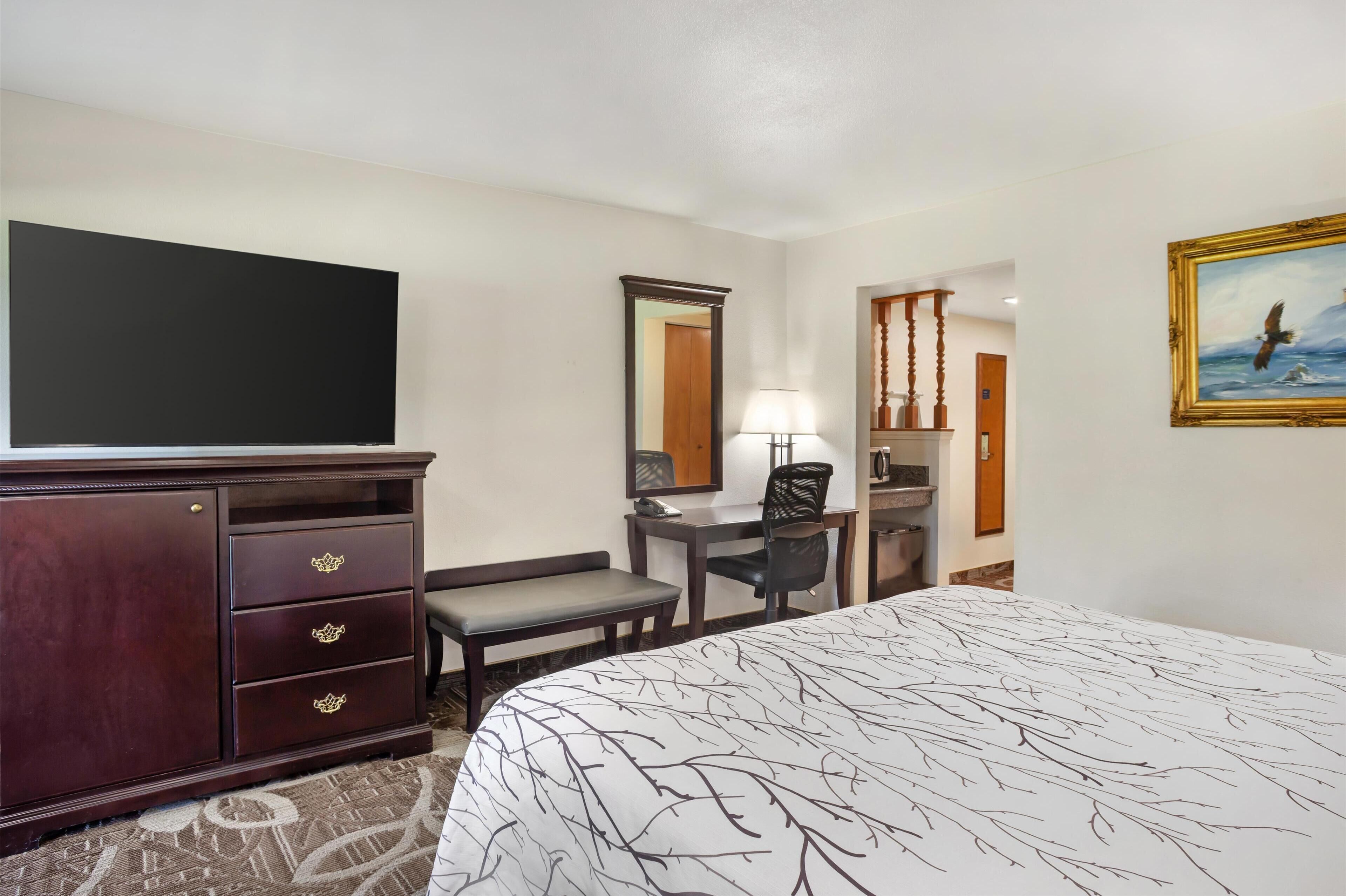 A hotel room with a large bed, a dresser with drawers, a desk with a chair, a mirror, a painting on the wall, and a television.