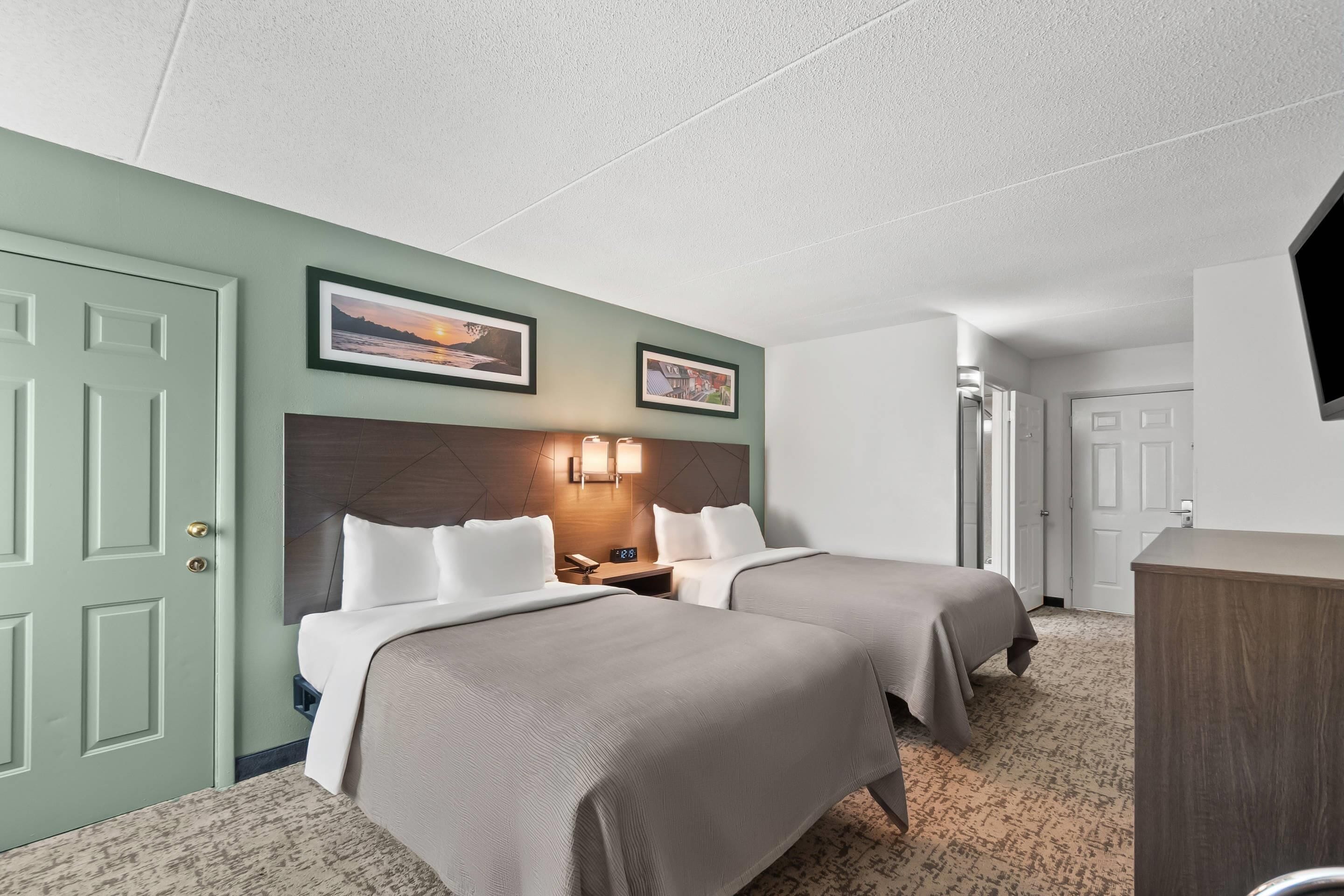 A hotel room with two beds, a wooden headboard, a wall-mounted TV, and a door to the right.