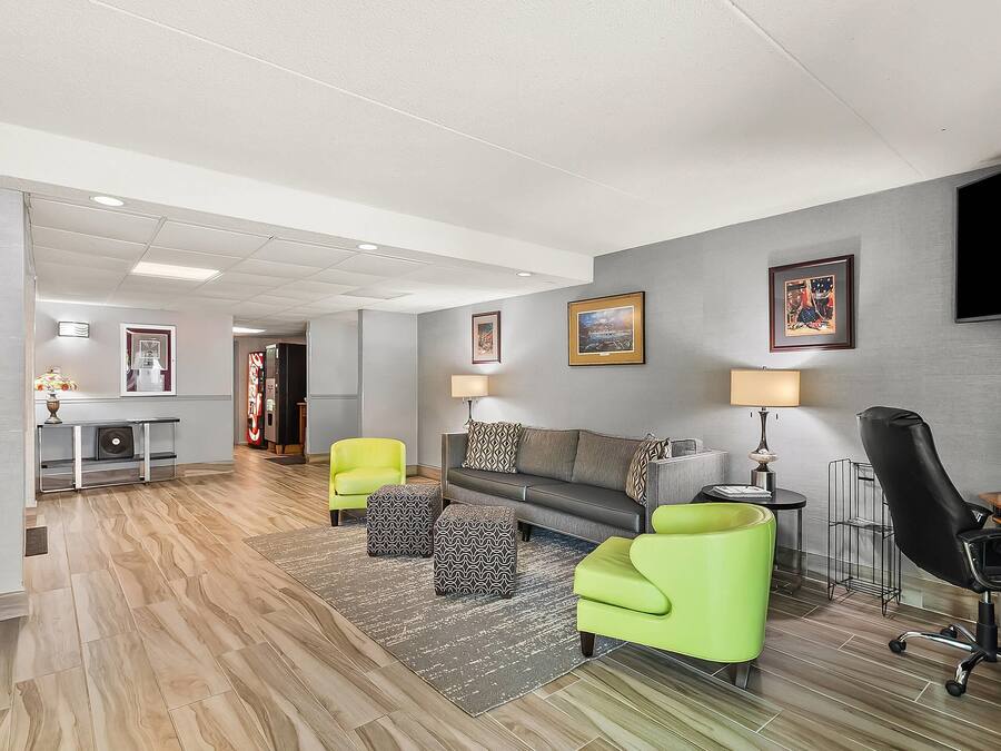 Uma sala de estar moderna com um sofá cinzento, poltronas verdes e uma cadeira de escritório preta. Há duas luminárias, uma televisão de tela plana e uma mesinha. O quarto possui piso de madeira e um tapete cinza.