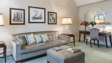 Un salon comprenant un canapé, un pouf et un bureau avec une chaise. Des tableaux encadrés ornent le mur et une lampe est posée sur le bureau.
