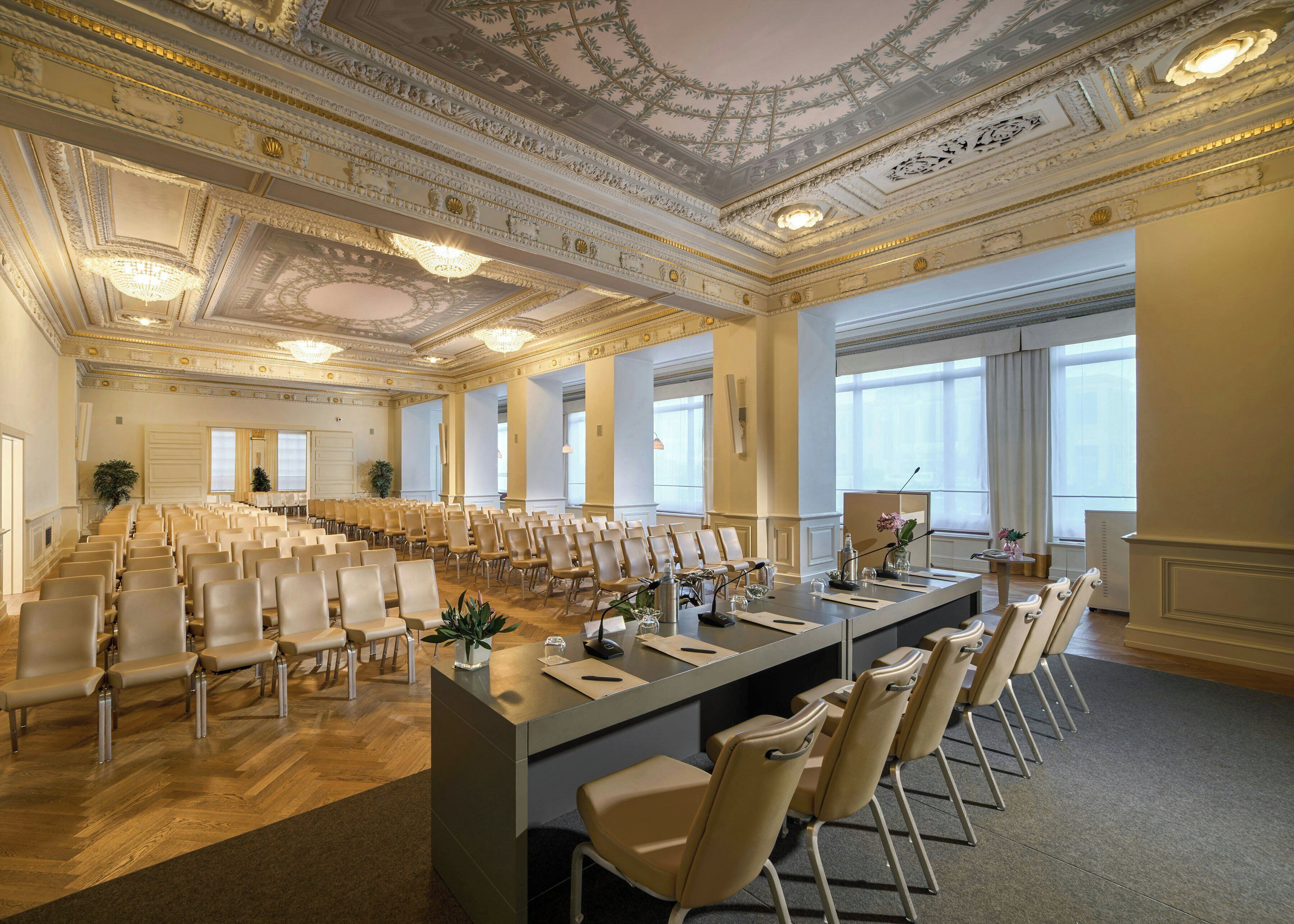 A conference room with rows of chairs, a long table with microphones, and a decorative ceiling.