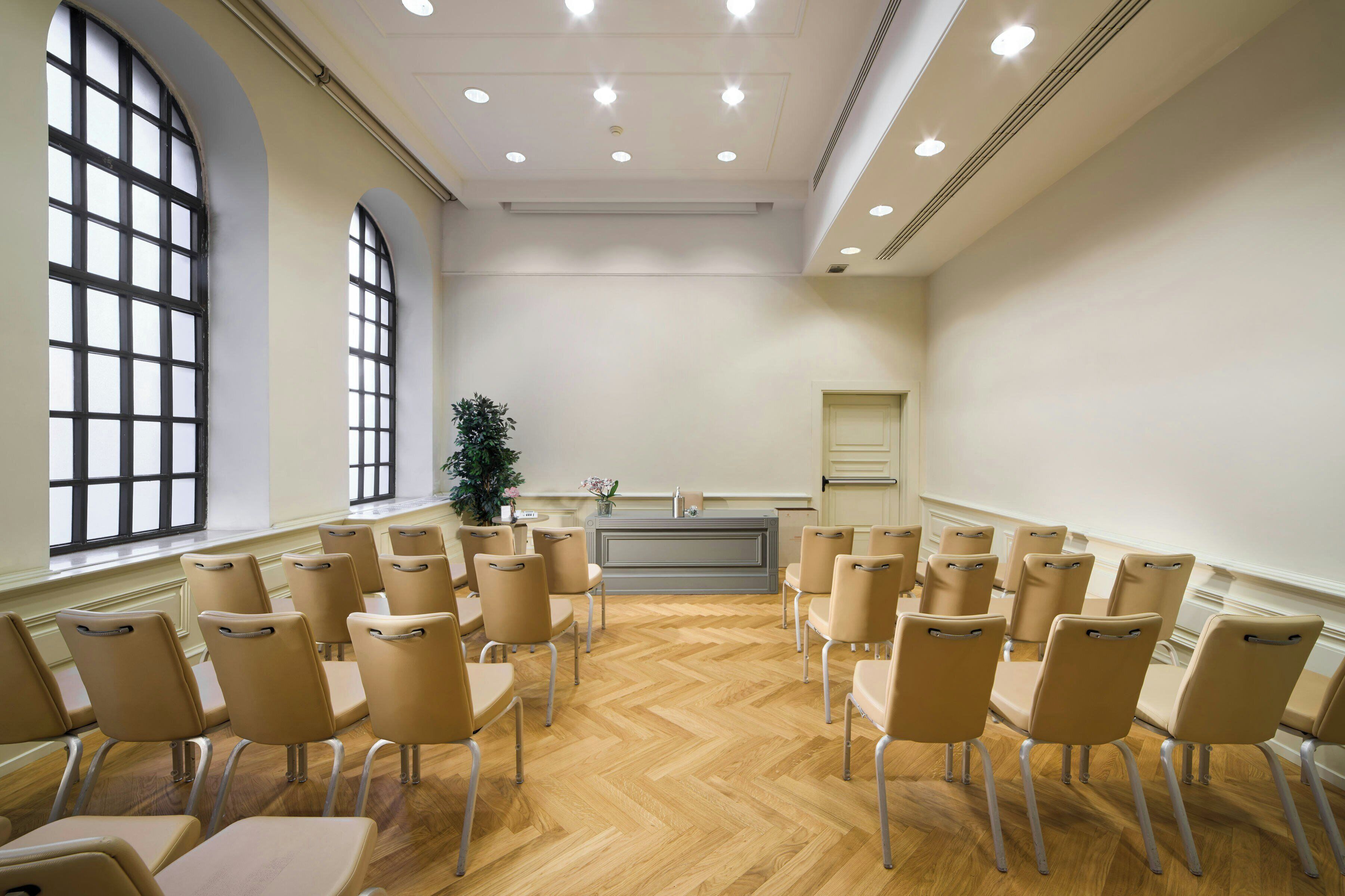 A conference room with beige chairs arranged in a semi-circle, a wooden floor, large windows, and a whiteboard.