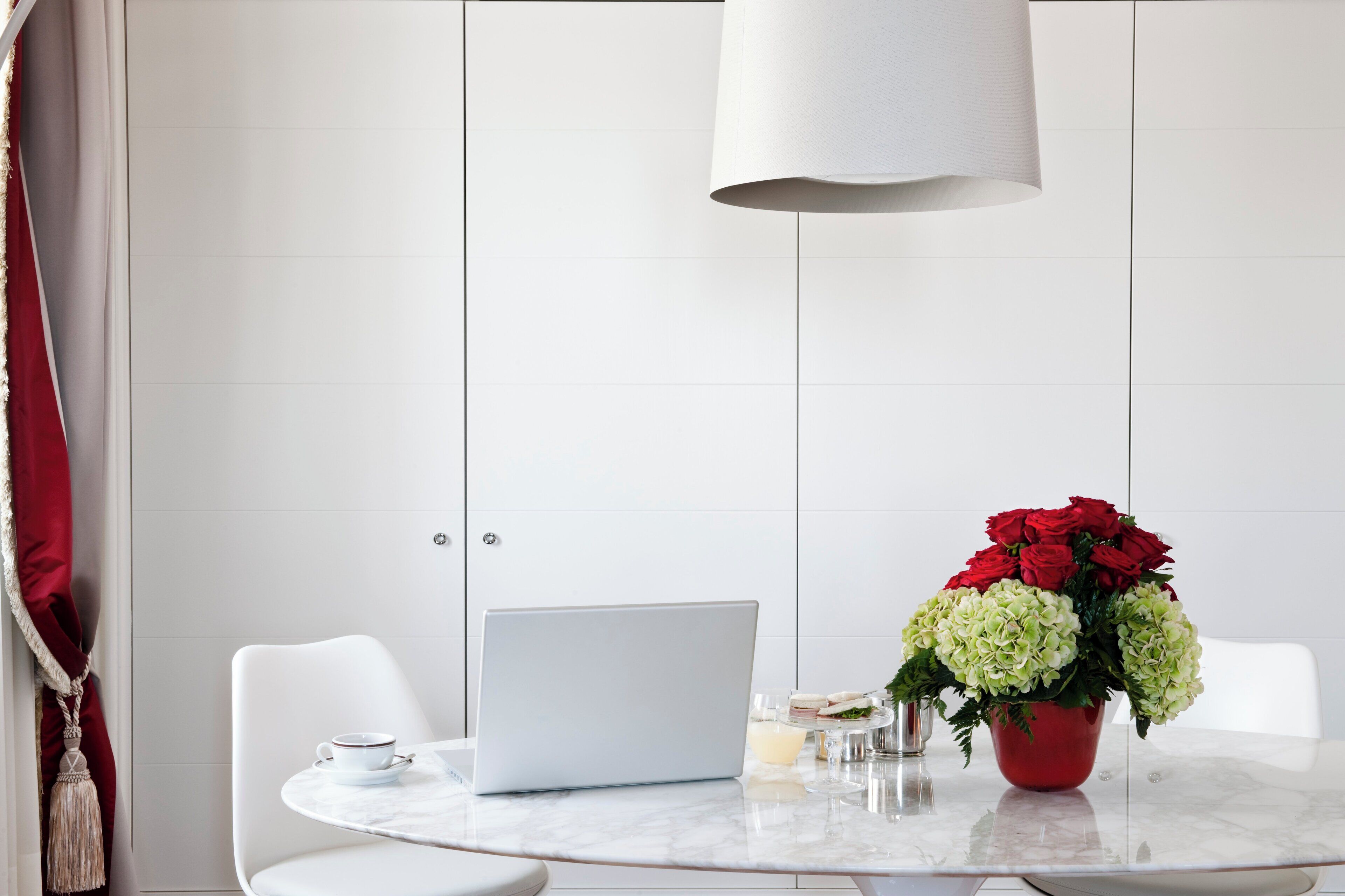 Ein moderner Essbereich mit einem weißen Tisch, einem Laptop, einer roten Vase mit Blumen und einem weißen Stuhl.