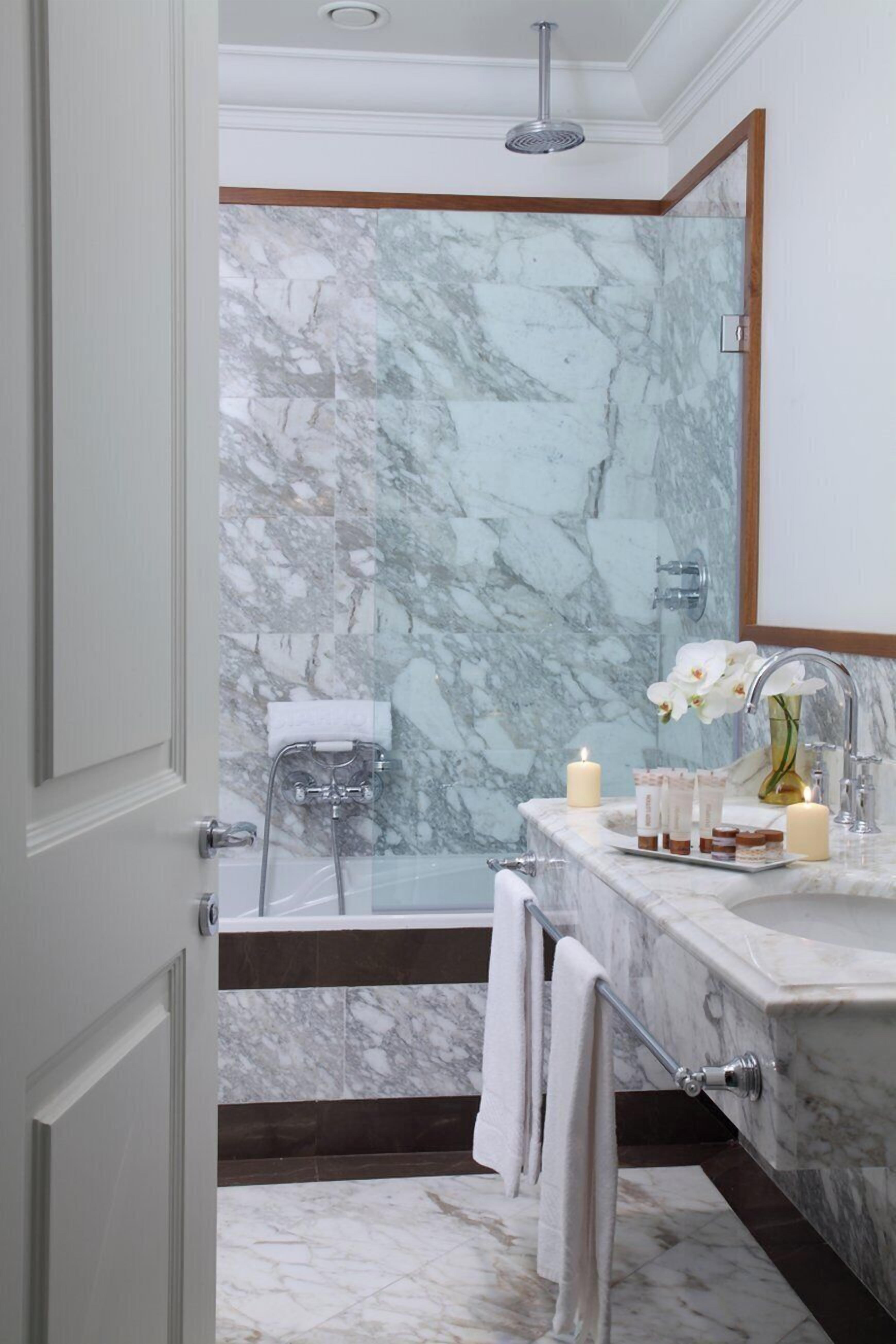 Un baño con pared de mármol, una bañera, un lavabo con espejo y un cabezal de ducha.