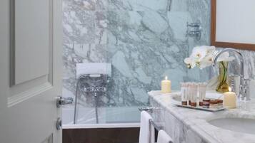 Un baño con pared de mármol, una bañera, un lavabo con espejo y un cabezal de ducha.