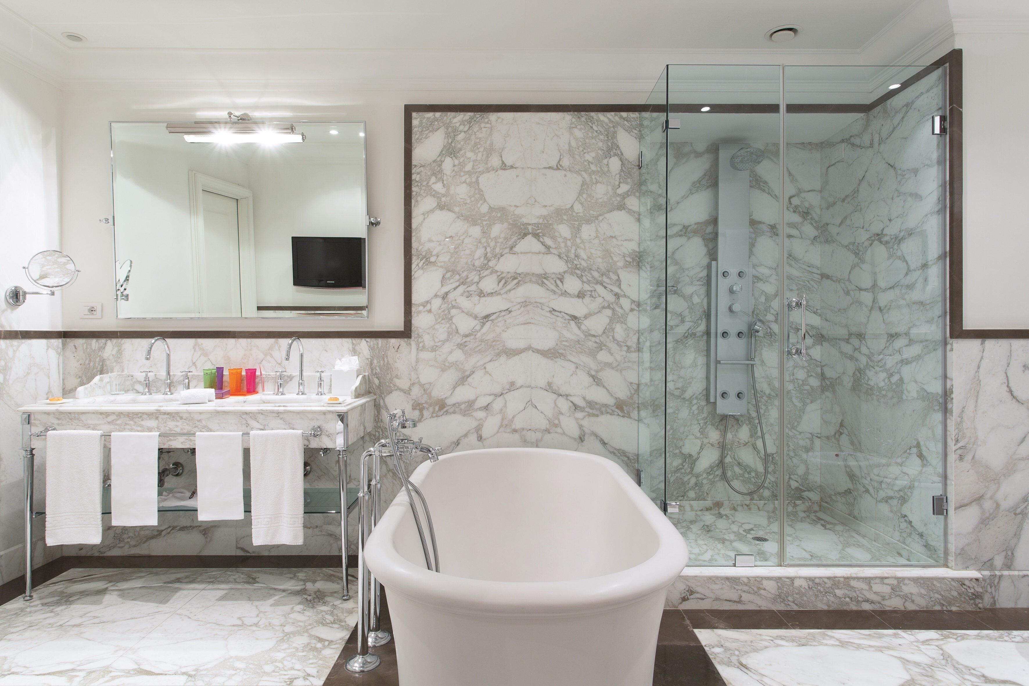 A modern bathroom with a glass-enclosed shower, a freestanding bathtub, and a vanity with a large mirror and a television.