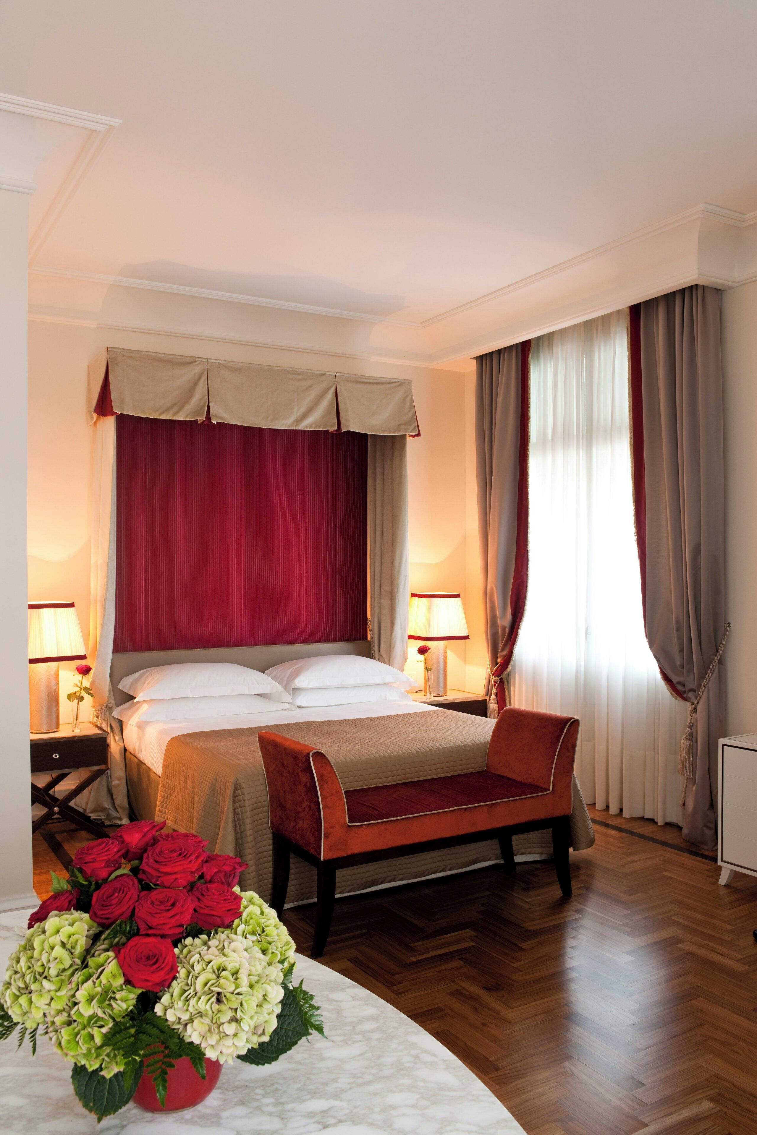 A hotel room with a bed, bedside tables, a bench, a vase of flowers, and curtains.