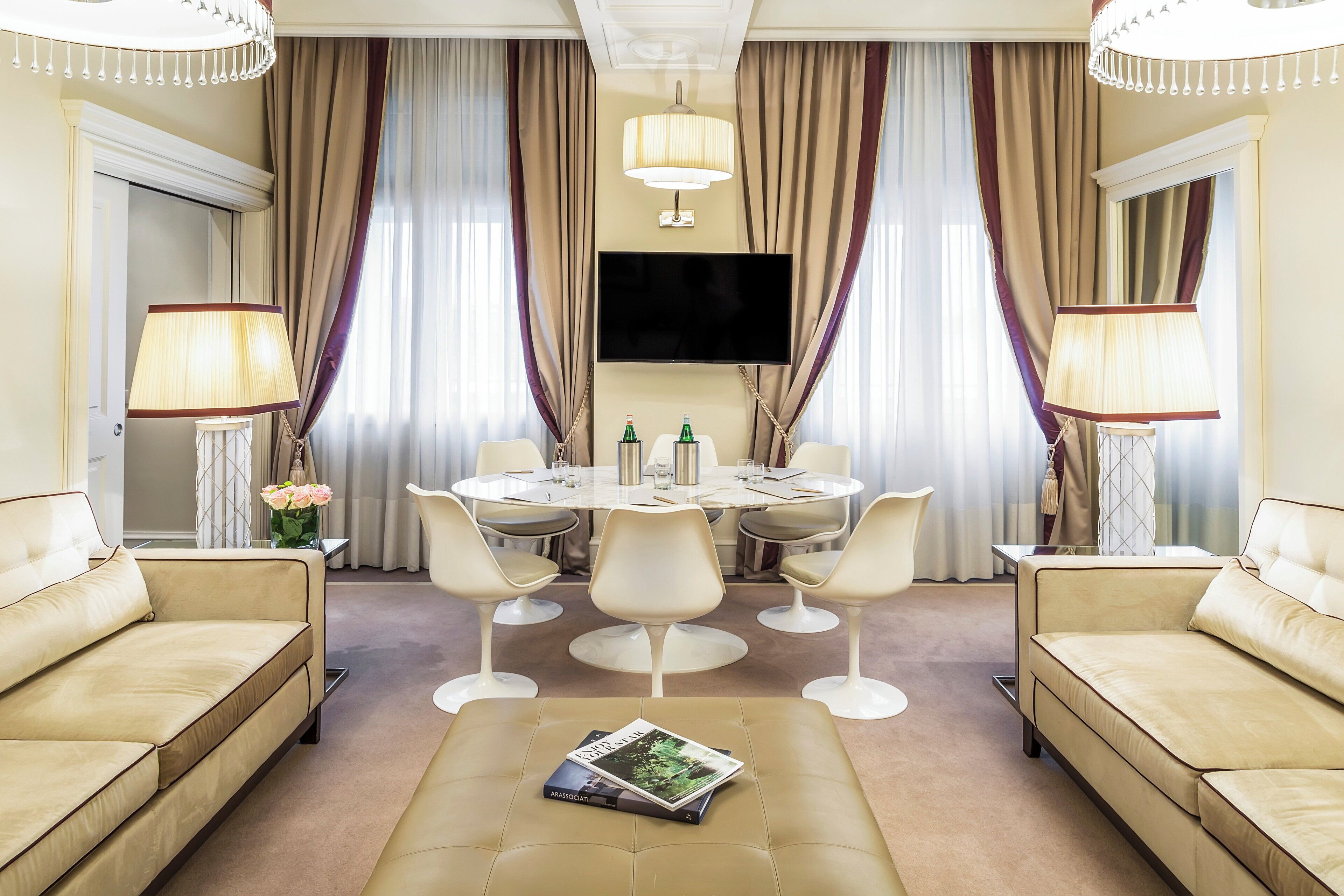 A hotel room with a beige sofa, a round dining table with chairs, a flat-screen TV, and a chandelier.
