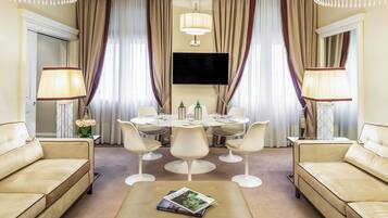 A hotel room with a beige sofa, a round dining table with chairs, a flat-screen TV, and a chandelier.