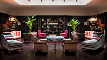 A spacious lounge with a high ceiling, dark wood flooring, and a central seating area with armchairs and a sofa. There are two large potted plants, a bookshelf filled with books, and a red and white striped sofa.