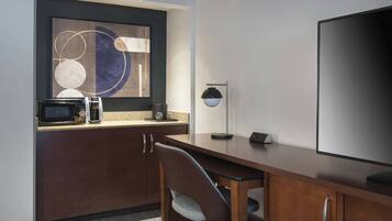 Habitación de hotel con escritorio de madera, una silla, televisión de pantalla plana, microondas, cafetera y una obra de arte abstracta enmarcada en la pared.