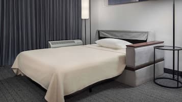 A hotel room with a bed, a bedside table, a lamp, an air conditioner, and a curtain.
