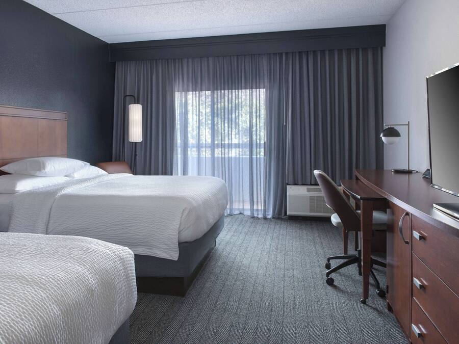 Une chambre d’hôtel avec deux lits, un bureau, une chaise, une télévision et une grande fenêtre avec des rideaux.