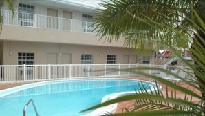 Ein Schwimmbecken mit weißem Zaun, einer Palme und einem Gebäude mit Balkon im Hintergrund.