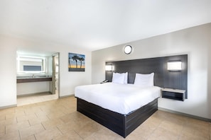 A hotel room with a large bed, a bedside table, a nightstand, a wall-mounted lamp, and a painting of palm trees.