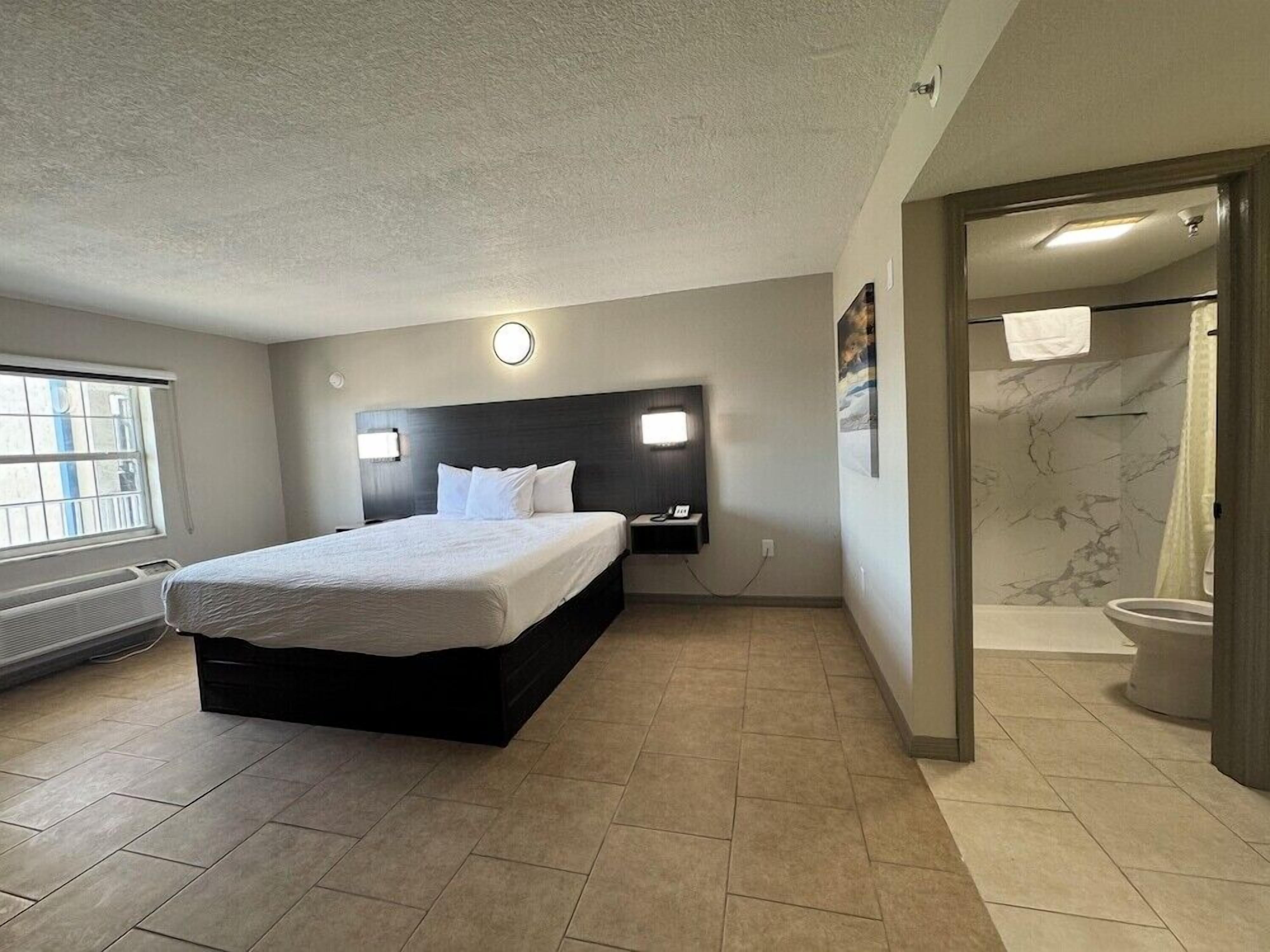Quarto de hotel moderno com uma cama grande, cabeceira com iluminação, banheiro com chuveiro e vaso sanitário, e janela com persianas.