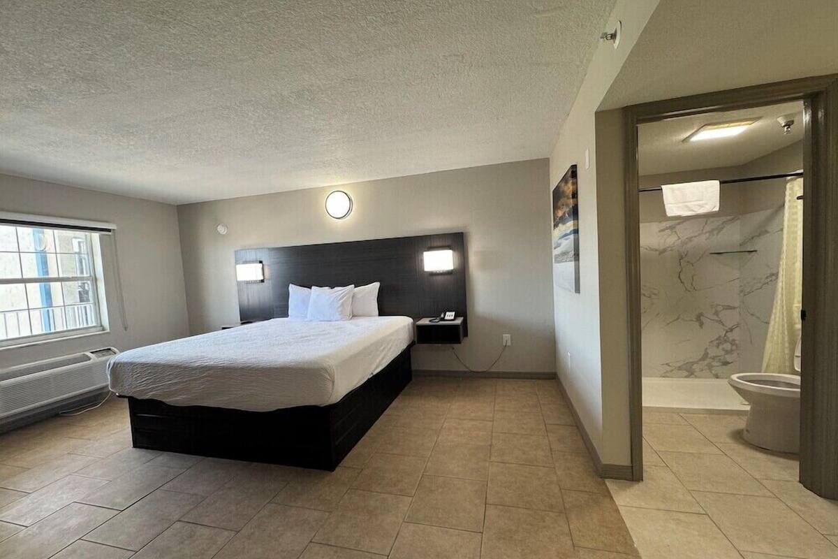 Een moderne hotelkamer met een groot bed, een hoofdbord met verlichting, een badkamer met douche en toilet, en een raam met luxaflex.