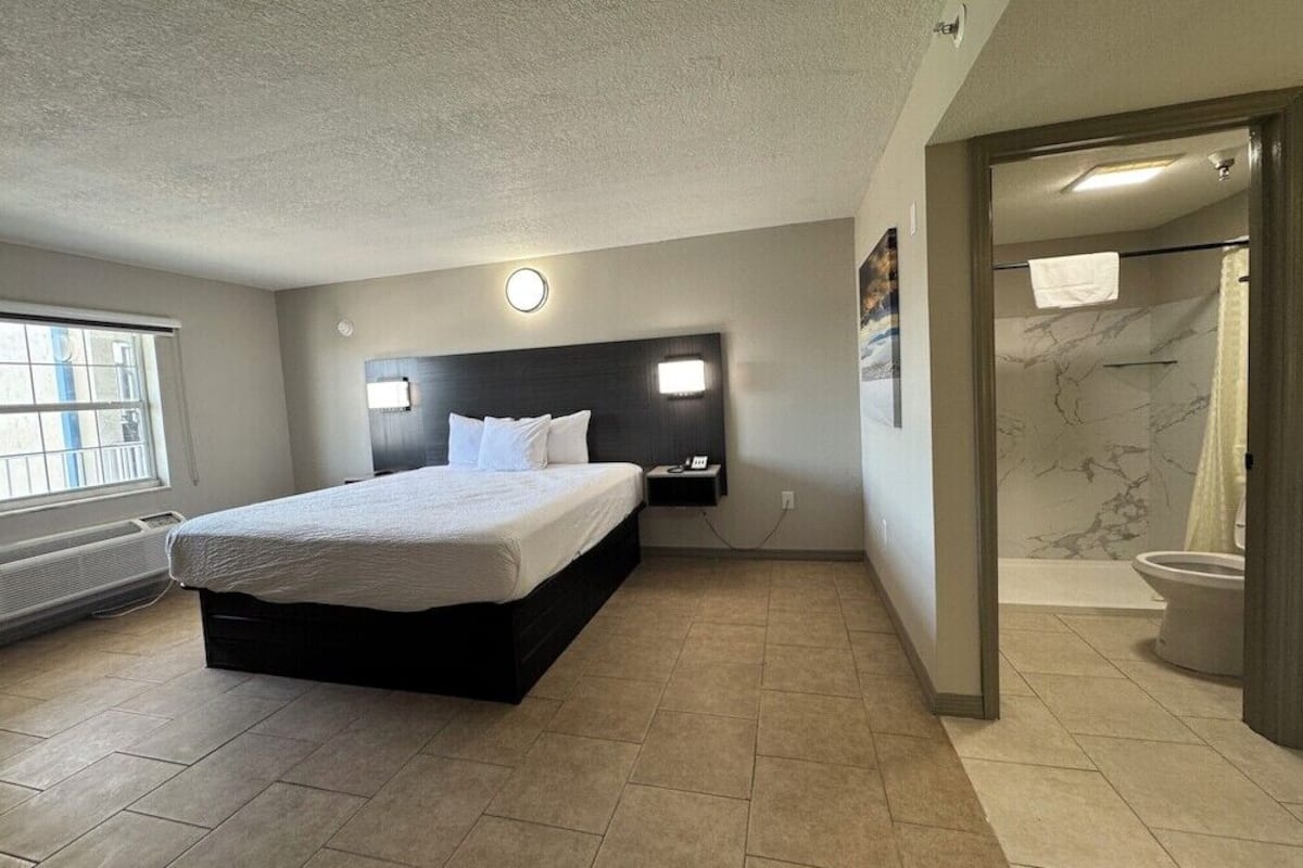 Quarto de hotel moderno com uma cama grande, cabeceira com iluminação, banheiro com chuveiro e vaso sanitário, e janela com persianas.