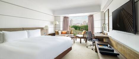 A hotel room with a large bed, a desk with a chair, a TV, and a view of the city.