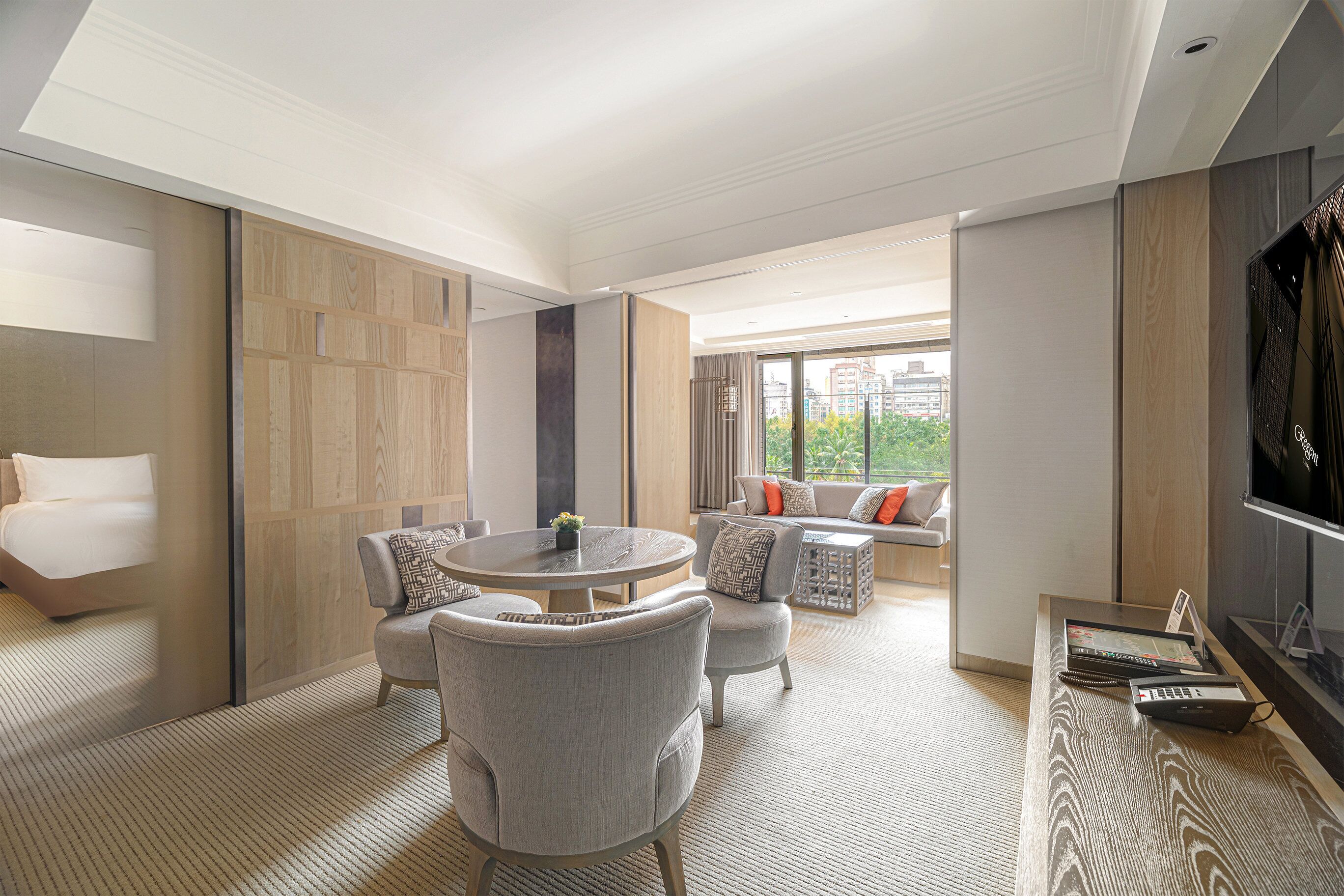 A modern hotel room with a large bed, a round dining table, two armchairs, a sofa, and a TV.