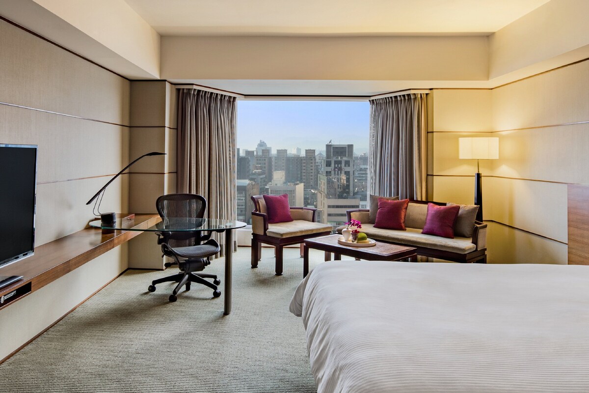 Habitación de hotel con cama, escritorio con silla, sofá, televisor y vista de la ciudad.
