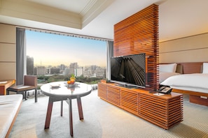 A hotel room with a large window, a round table with fruit, a TV, and a bed.