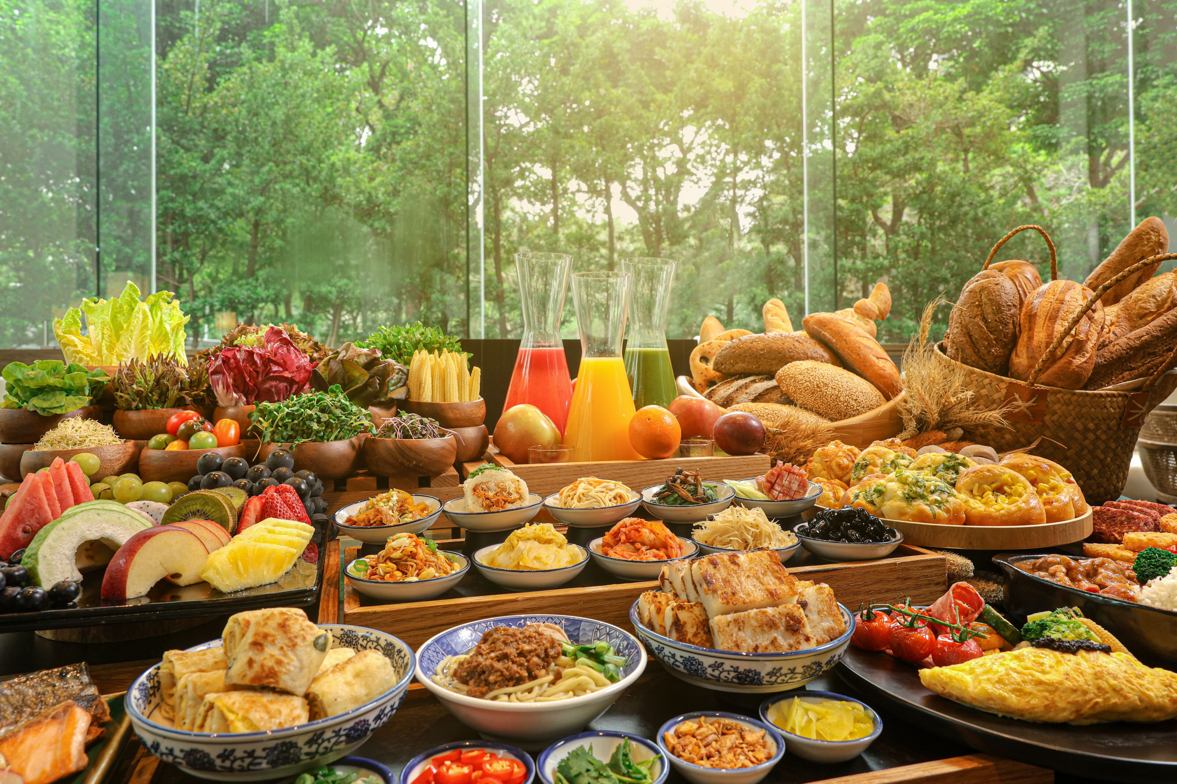 A table with a variety of dishes and beverages, set against a backdrop of a forest view through large windows.