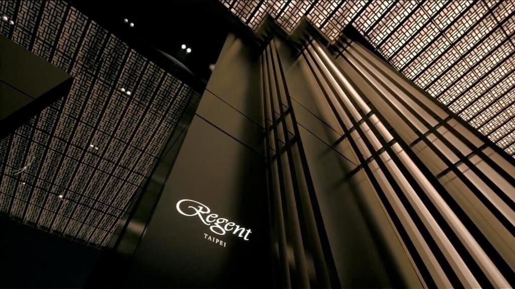A modern interior with a prominent 'Regent Taipei' sign, featuring a geometric ceiling design and vertical structural elements.