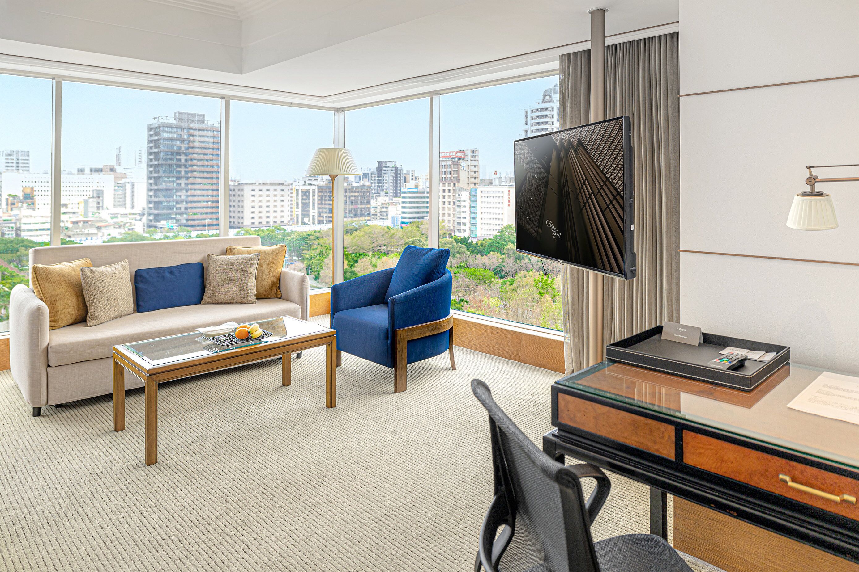 Habitación de hotel con sofá, sillón, escritorio y un amplio ventanal con vistas a la ciudad.