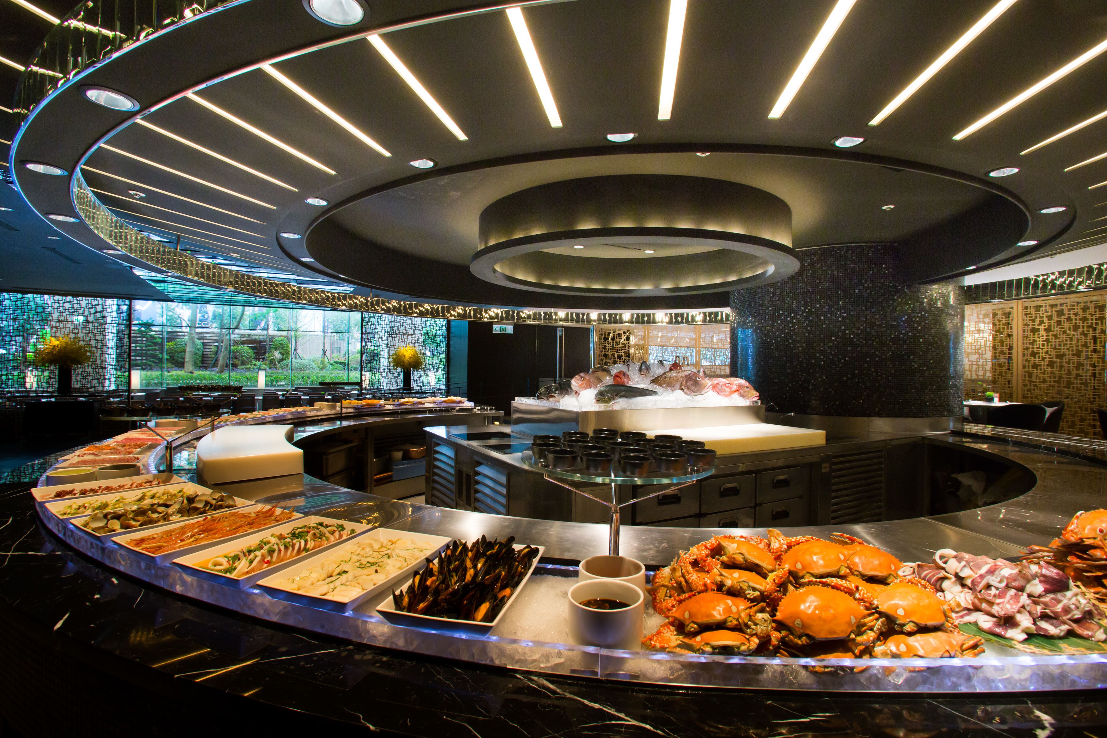 A buffet with seafood and various dishes, a modern ceiling design, and a view of the outdoors.