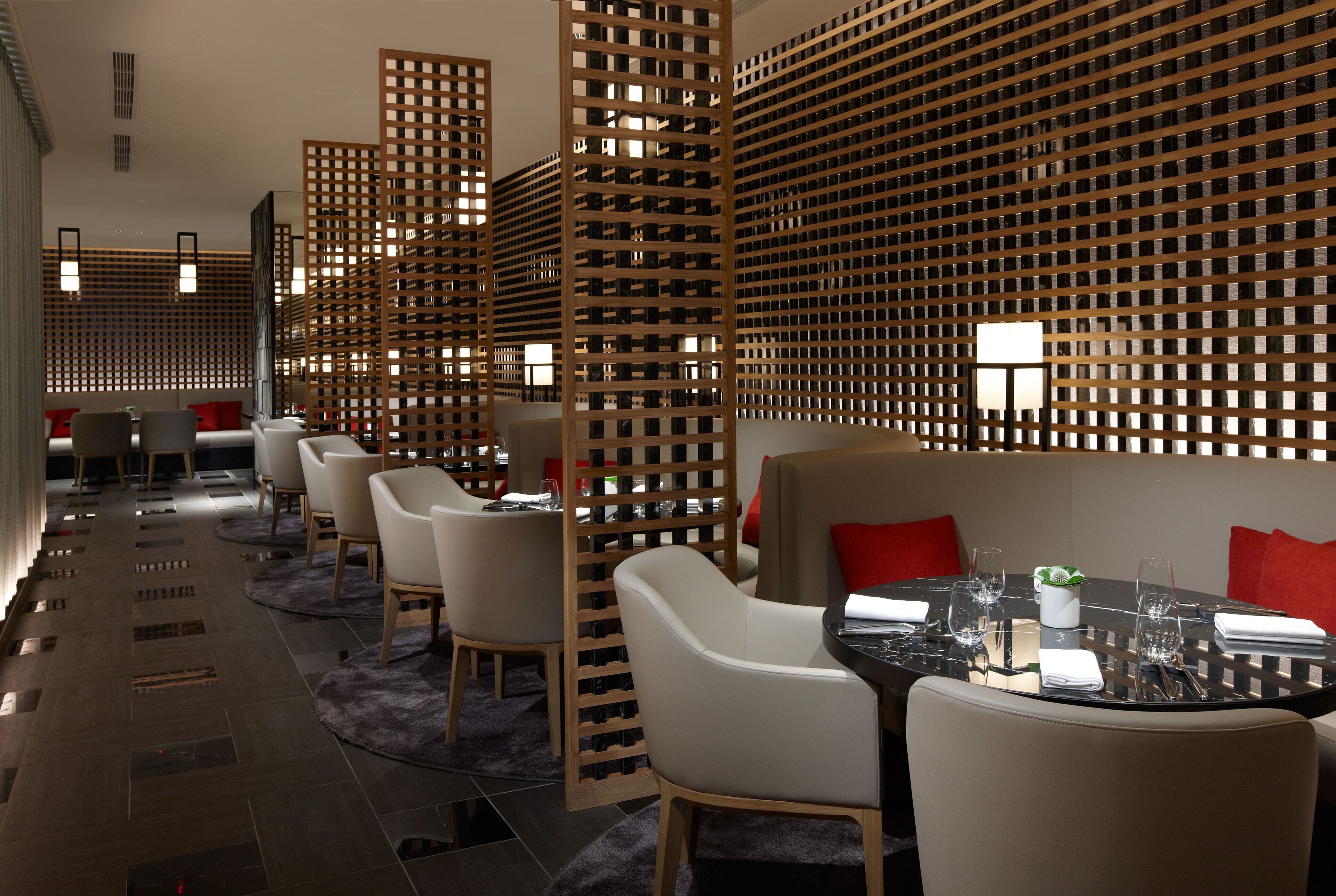 Un restaurant moderne avec de hauts plafonds, des cloisons en treillis de bois et une grande table à manger centrale, agrémentée de chaises et de coussins rouges.