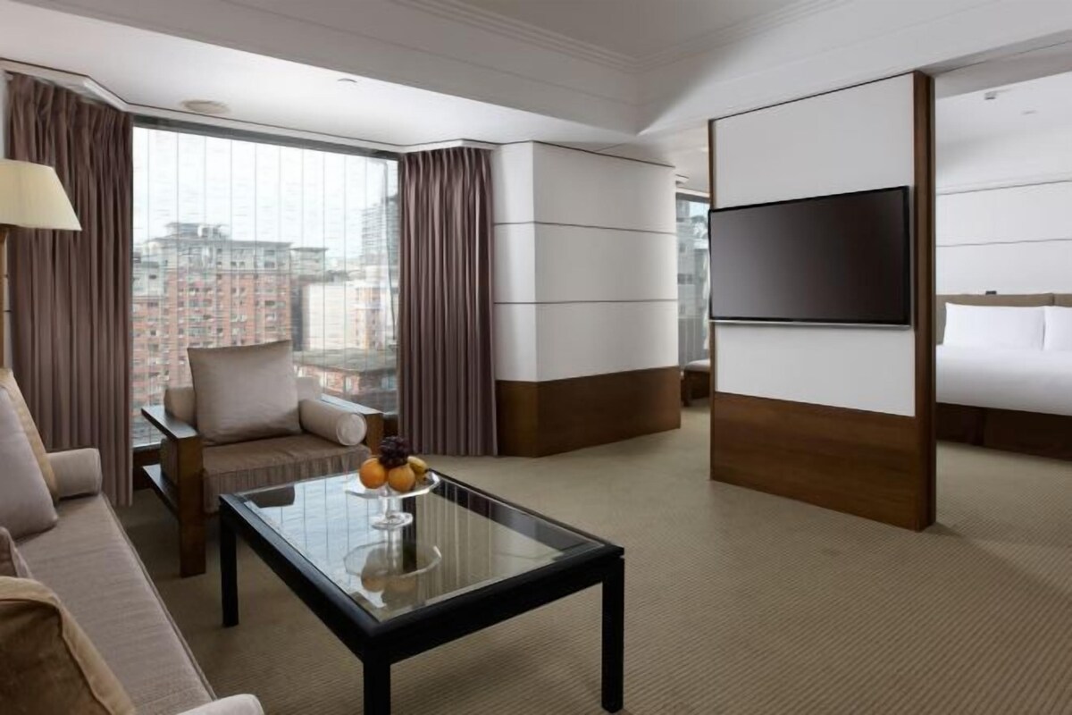Habitación de hotel con un ventanal grande, un sofá, una mesa de centro con fruta, un televisor y una cama.