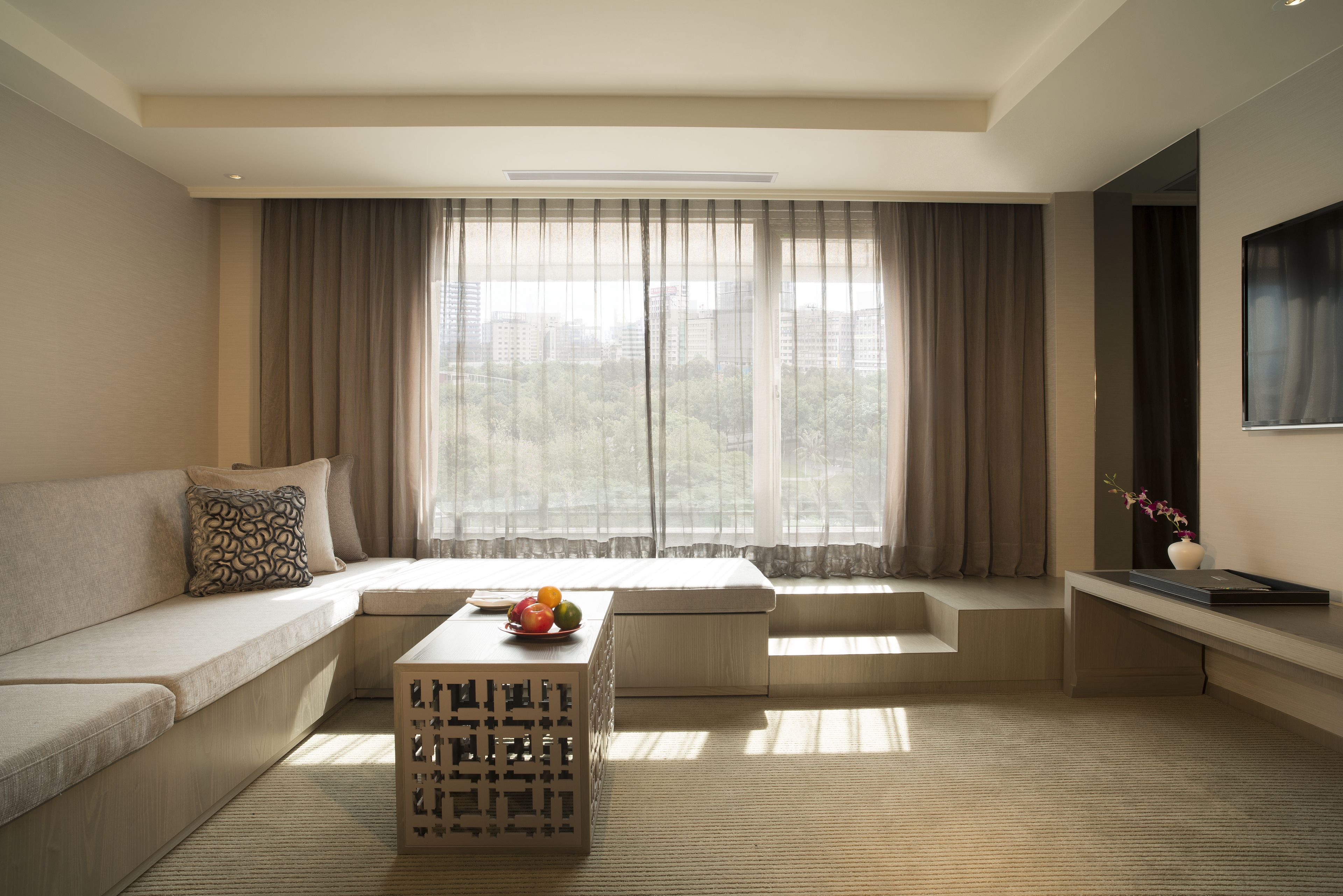A modern living room with a sofa, a coffee table with fruit, a small table with a vase, and a large window with curtains.