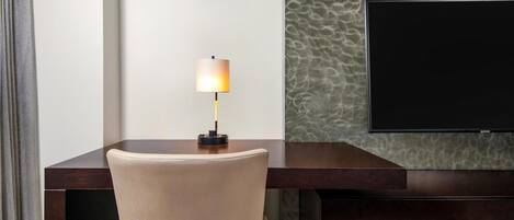 Een hotelkamer met een bureau, een stoel, een lamp en een flatscreen televisie die aan de muur is bevestigd.