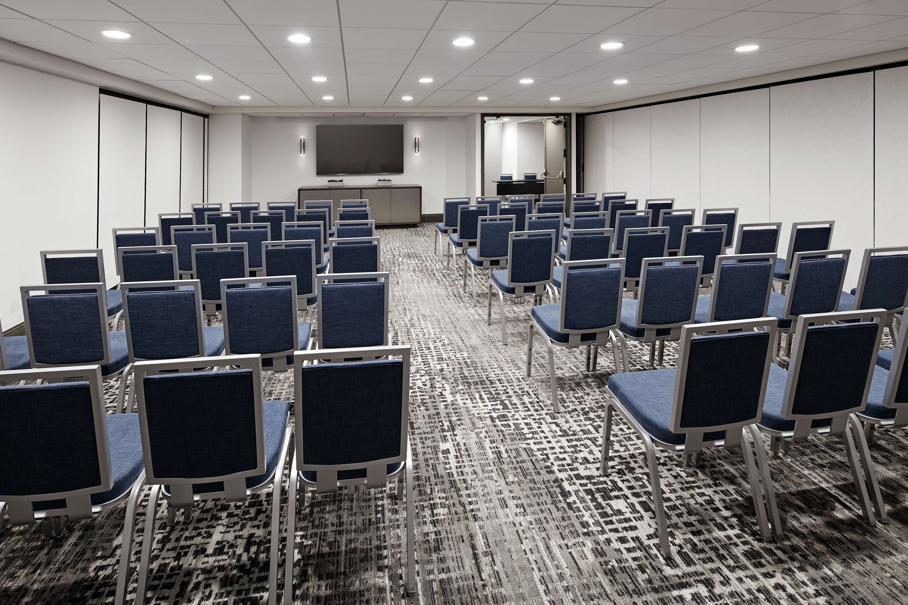 Uma sala de conferência com fileiras de cadeiras, uma tela grande e piso carpeteado.