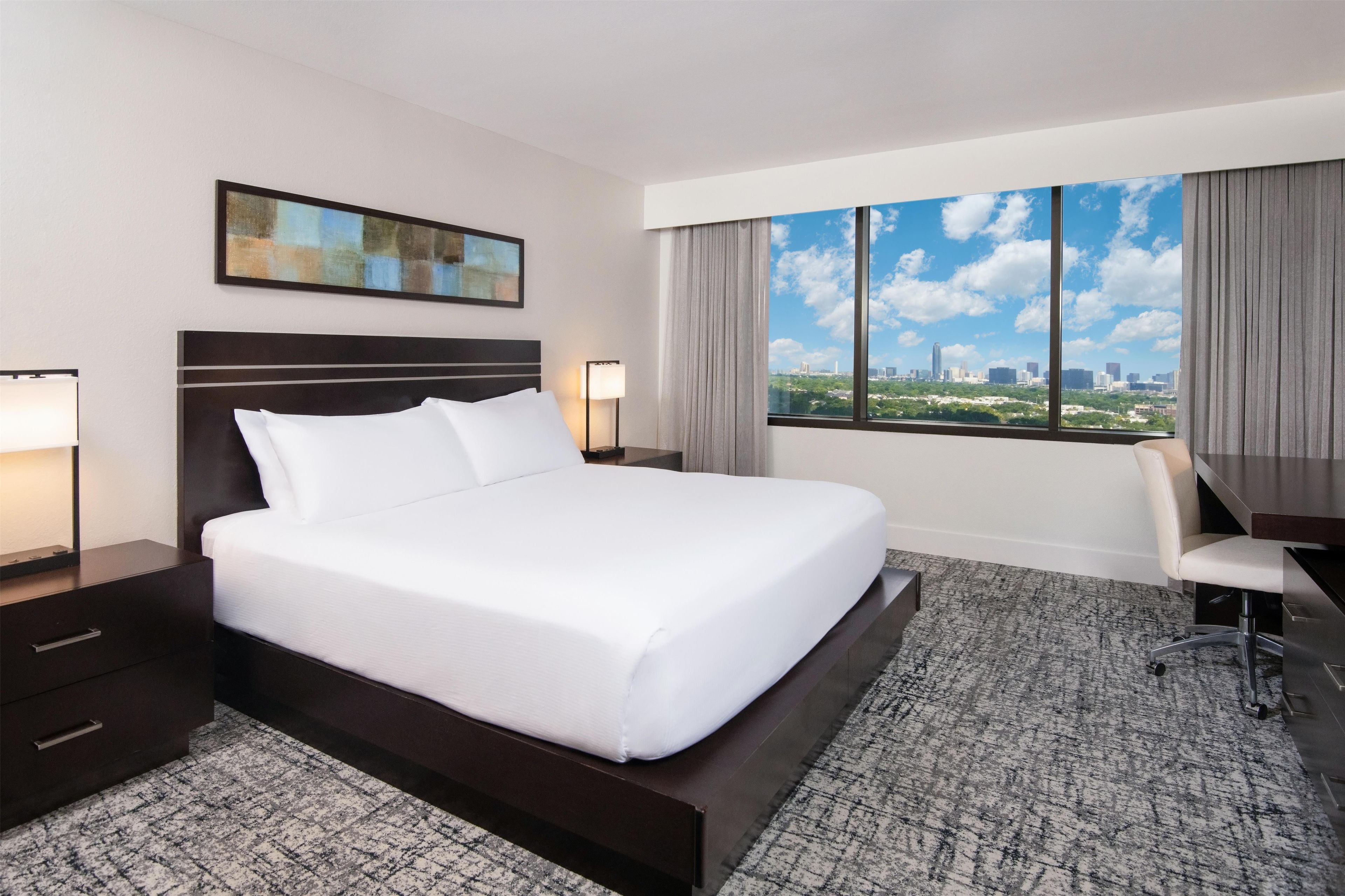 Una habitación de hotel con una cama grande, un escritorio, una silla, una mesita de noche, una lámpara, un cuadro en la pared y una ventana con vistas a la ciudad.