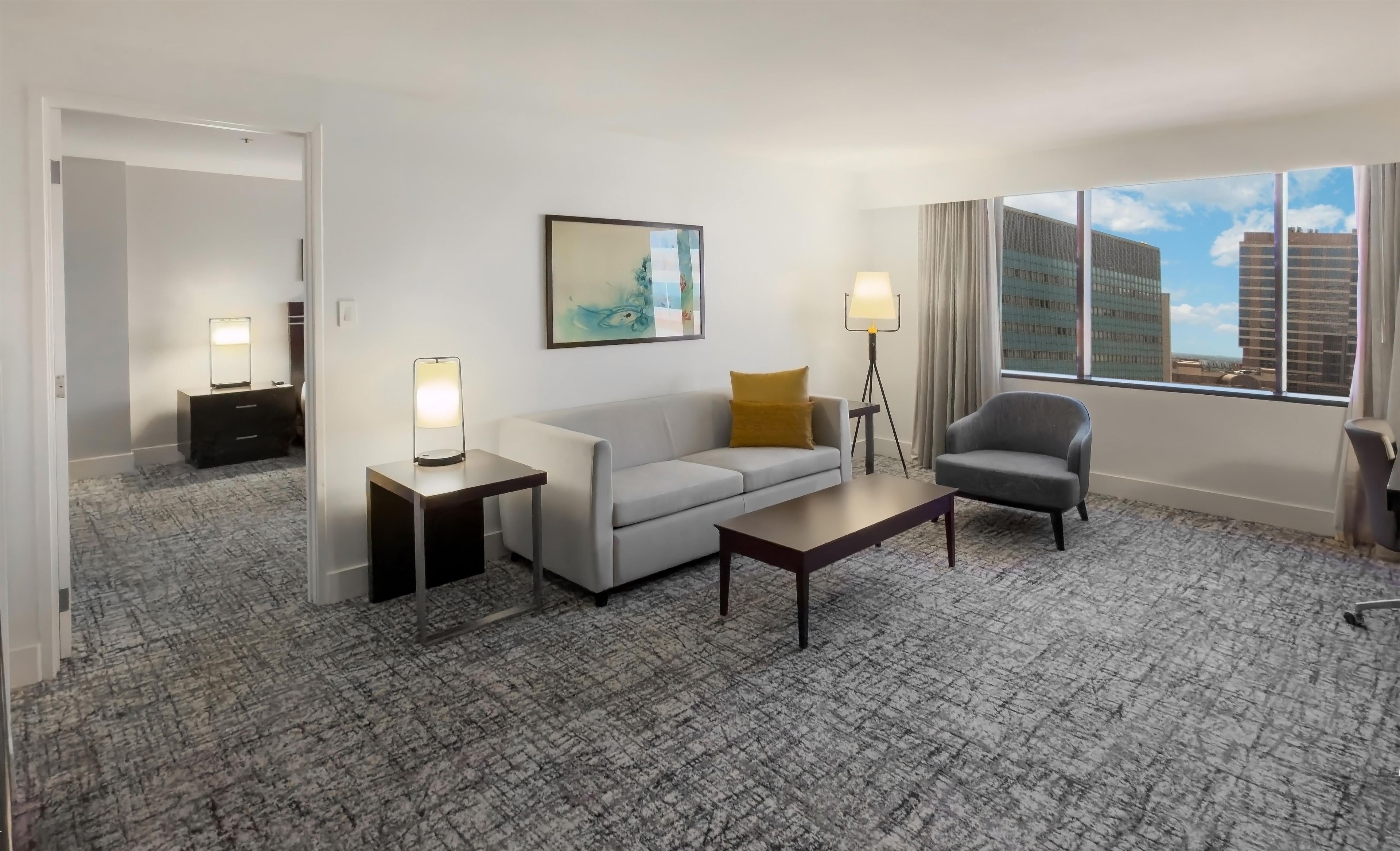 A hotel room with a sofa, armchair, side table, lamp, and a painting on the wall.