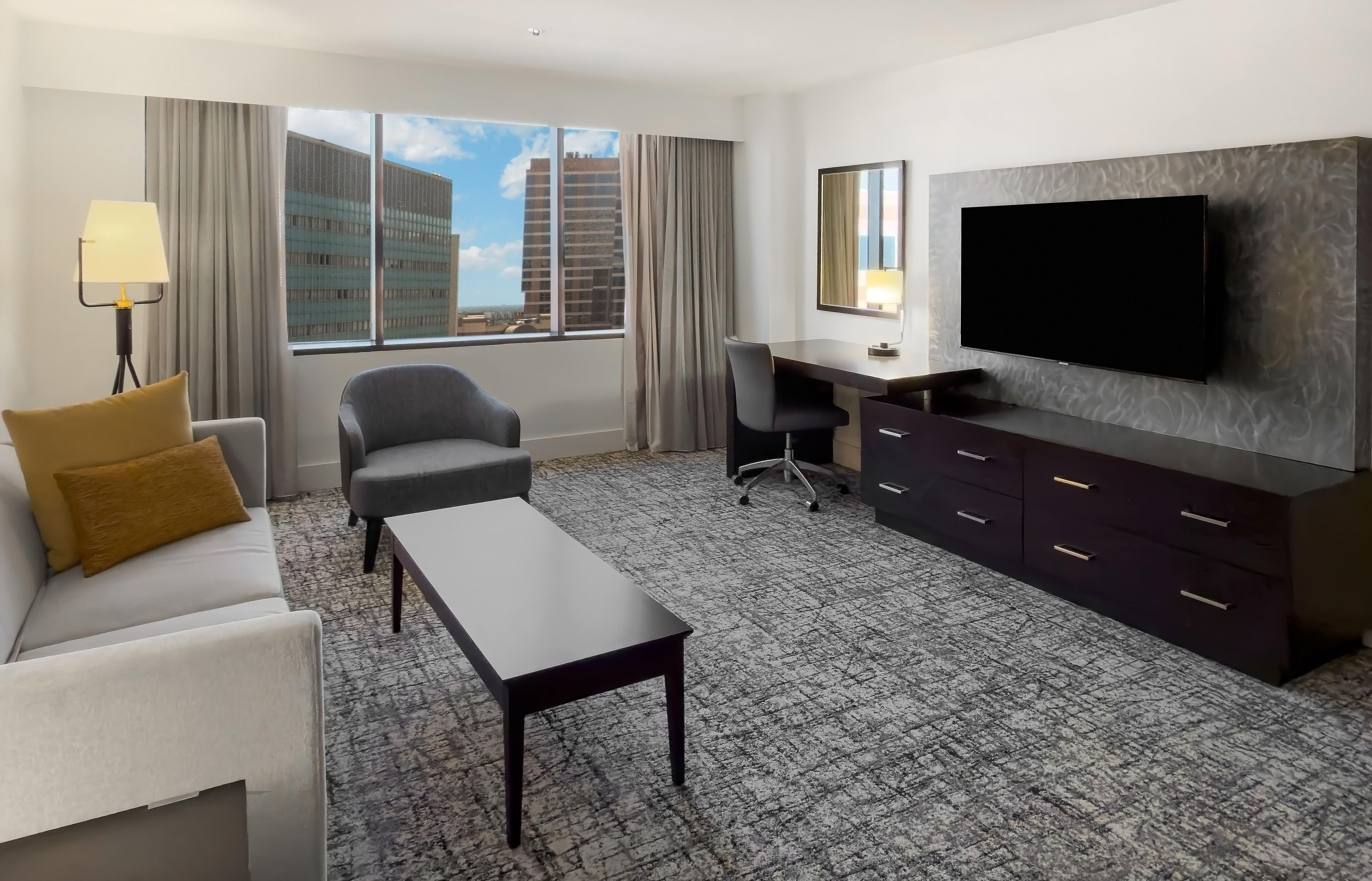 A hotel room with a sofa, a desk, a chair, a television, and a large window with a city view.