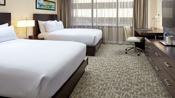 A hotel room with two beds, a desk, a chair, a TV, and a large window with curtains. - Hilton Houston Plaza/Medical Center (Houston)