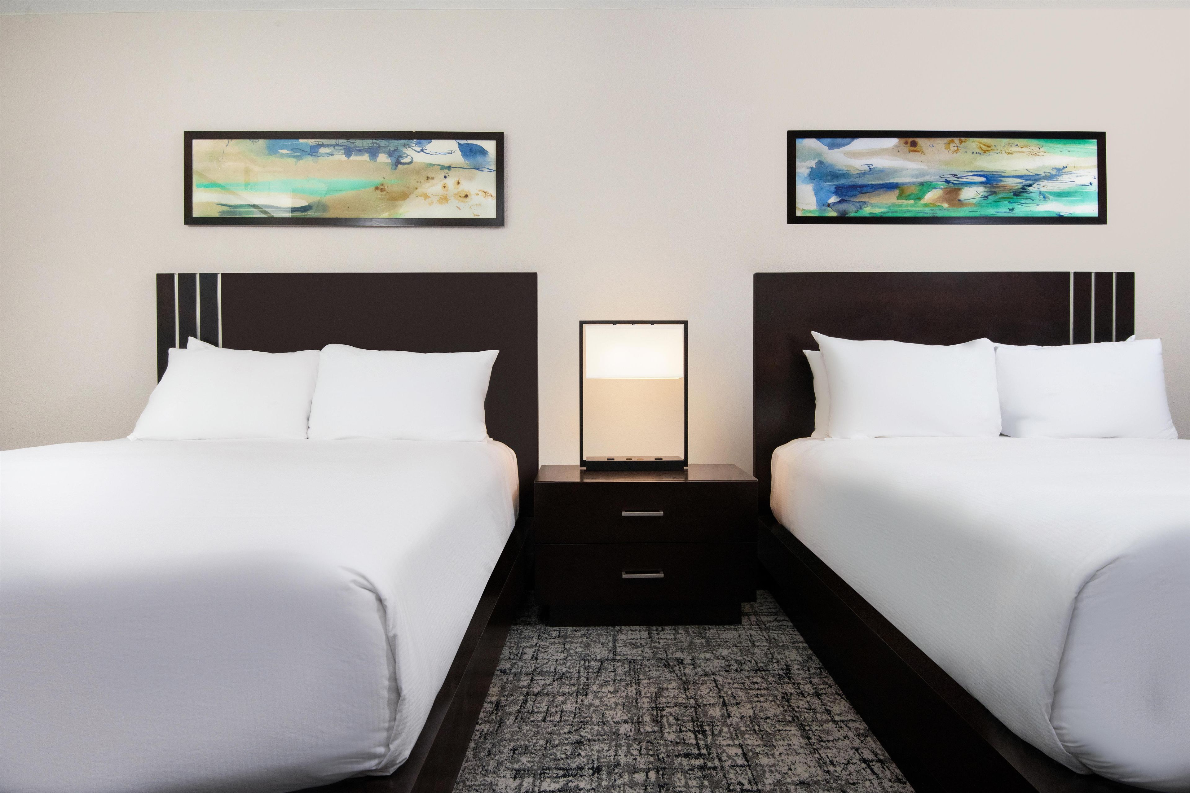 Una habitación de hotel con dos camas, una mesita de noche con lámpara y dos cuadros en la pared.