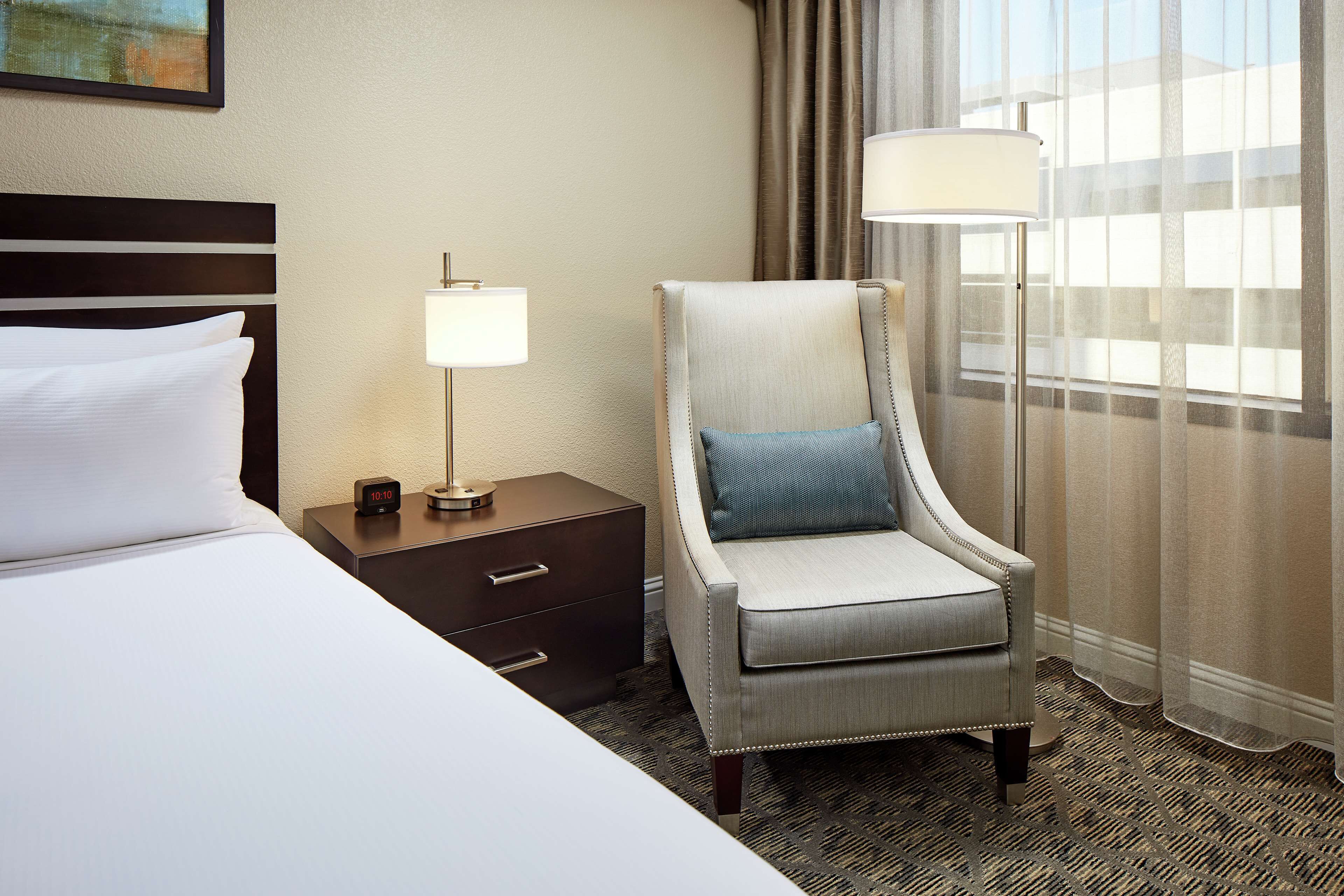 Quarto de hotel com cama, mesa de cabeceira com abajur, cadeira e janela com cortinas.