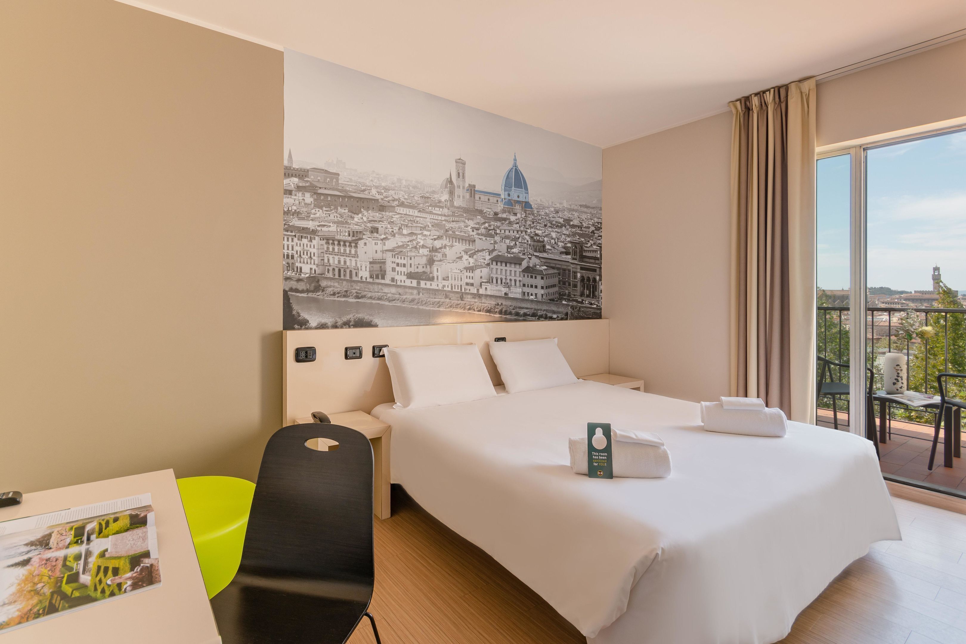 Habitació d'hotel amb un llit gran, una taula de treball amb una cadira, una finestra amb vista a la ciutat i un balcó amb una cadira.