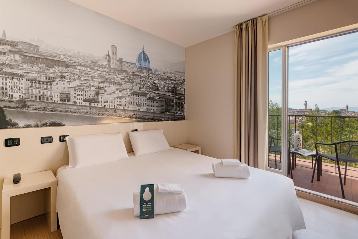 Hotelkamer met een groot bed, een nachtkastje, een hangend horloge en uitzicht op een stadsgezicht vanaf een balkon.