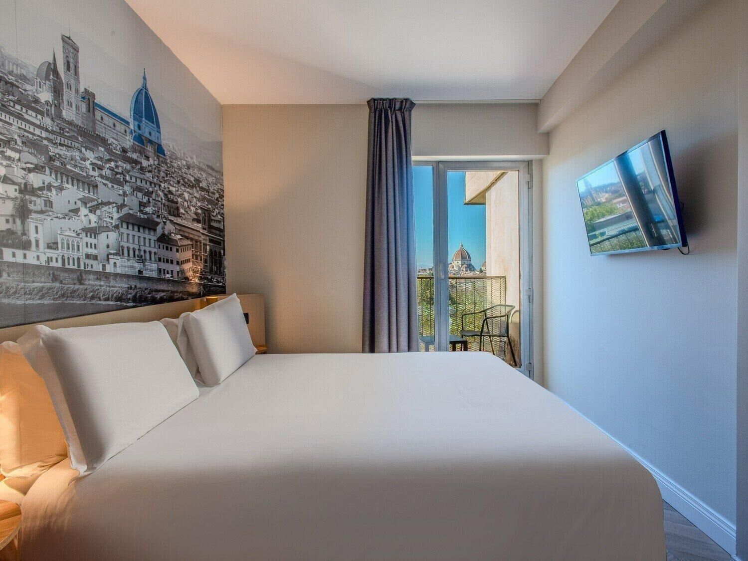 Habitación de hotel con una cama grande, un mural con vista a la ciudad, un televisor y un balcón con vistas a una cúpula y edificios.