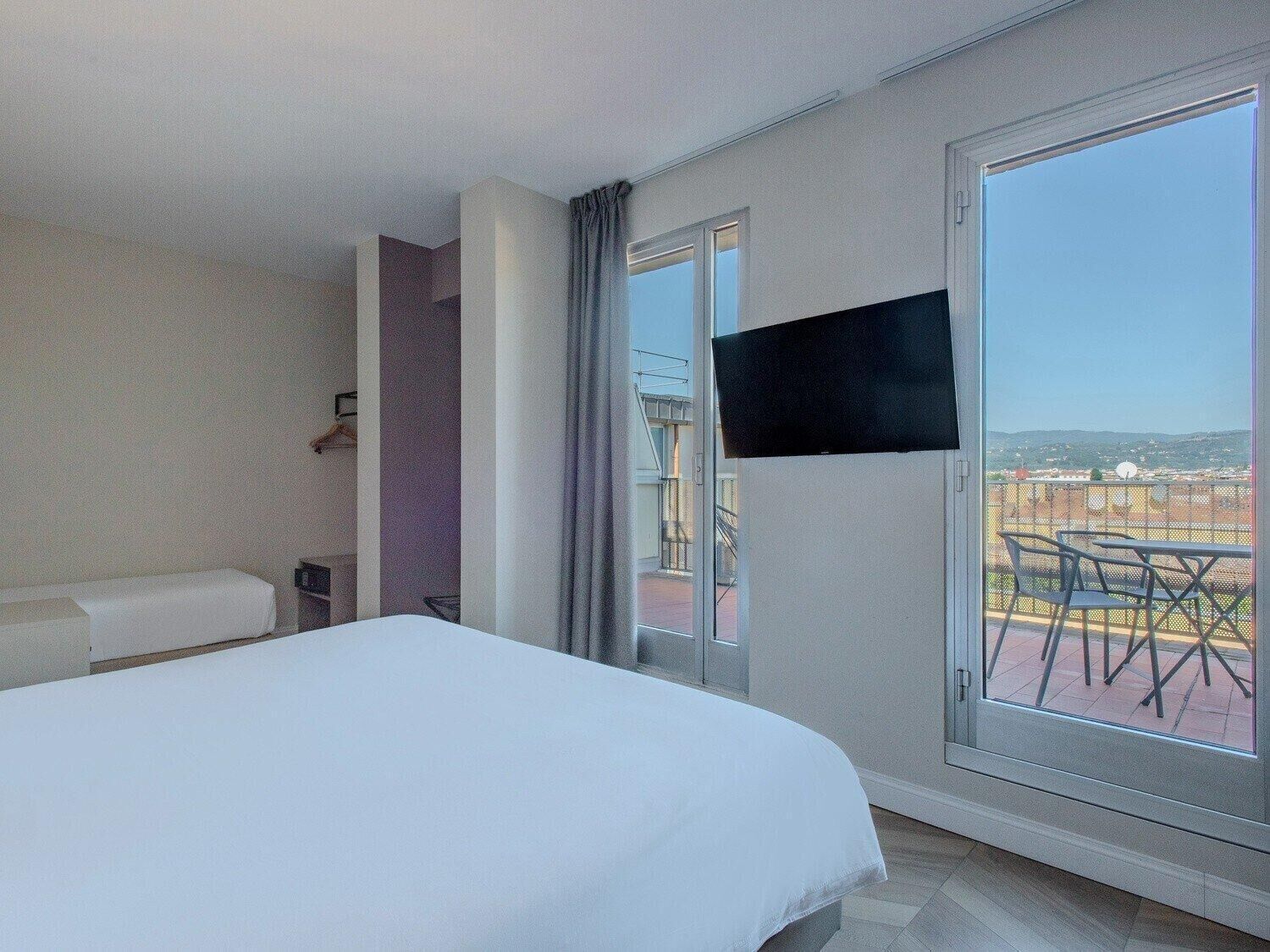 Habitación de hotel con cama, televisor montado en la pared y balcón con vistas a la ciudad.